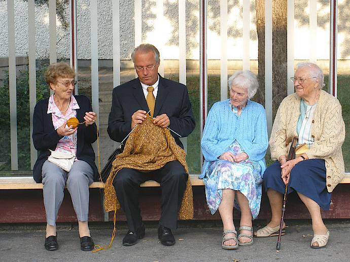Entraide : Homme tricotant entouré de vieilles dames qui l'aident.